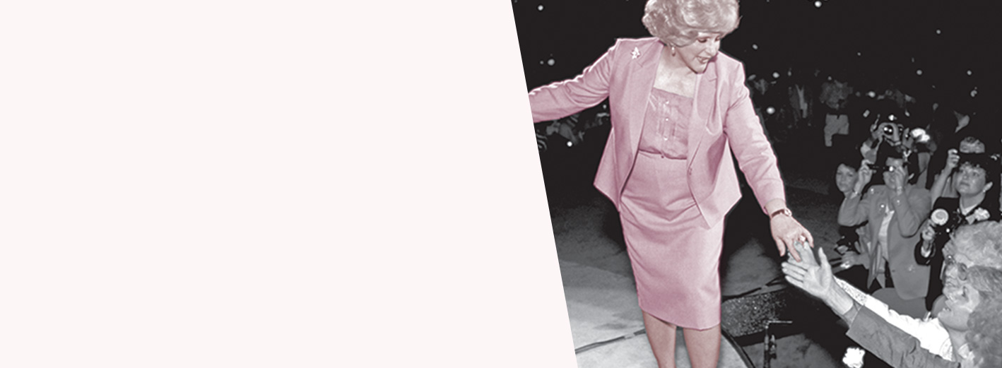 Mary Kay Ash está em cima de um palco e estende a mão para as consultoras da Força de Vendas Independente da Mary Kay na platéia.