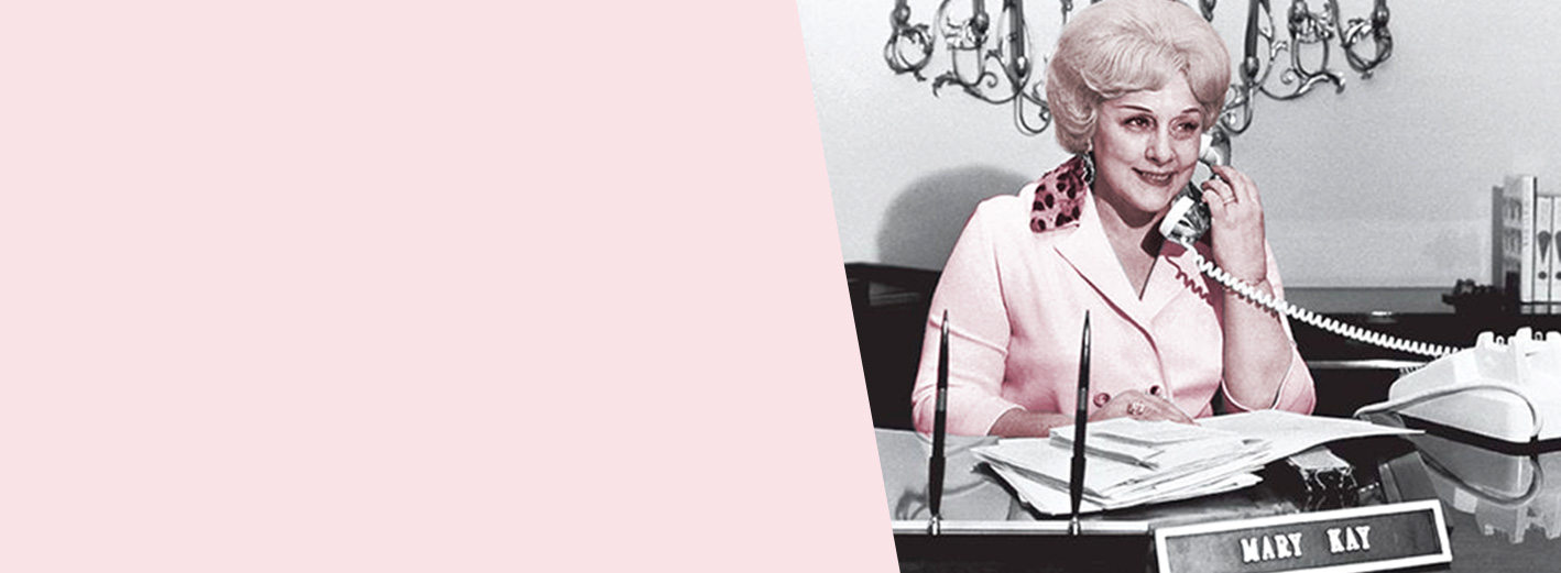 Uma Mary Kay Ash jovem sorri enquanto faz um telefonema de sua mesa.