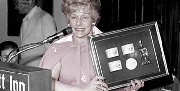 Mary Kay Ash sorri para uma foto enquanto sobe em um pódio e segura uma placa.